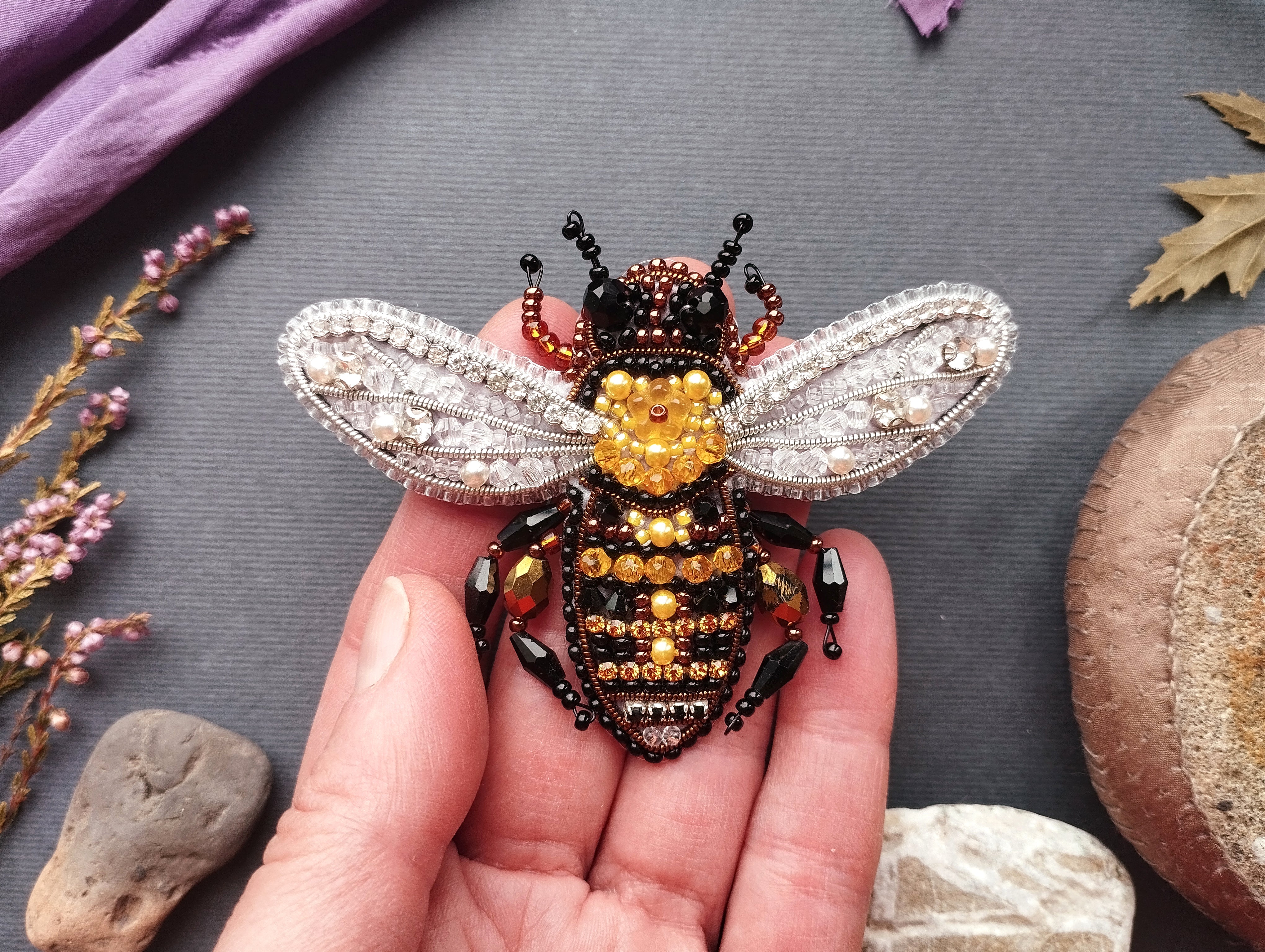 A hand holding a decorative beaded bee brooch against a gray background.